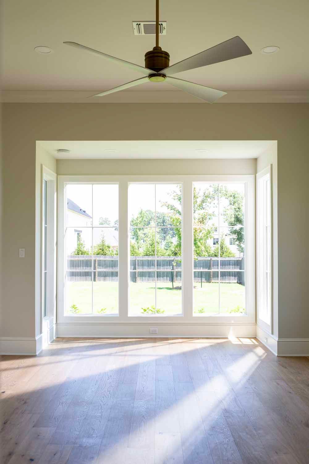 A bright, minimalist room with light wood floors, a modern ceiling fan, and large windows overlooking a backyard.