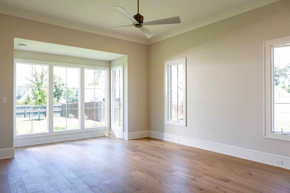 Empty room with light-colored walls, wood floors, a ceiling fan, and large windows looking out onto a backyard.