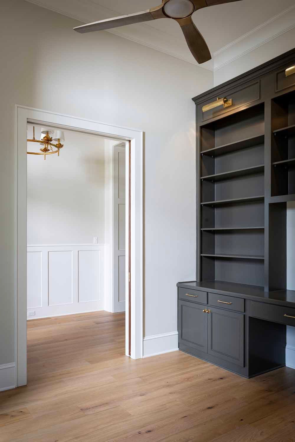 A view through a doorway into a room with white wainscoting, next to a dark gray built-in bookshelf with brass lights.