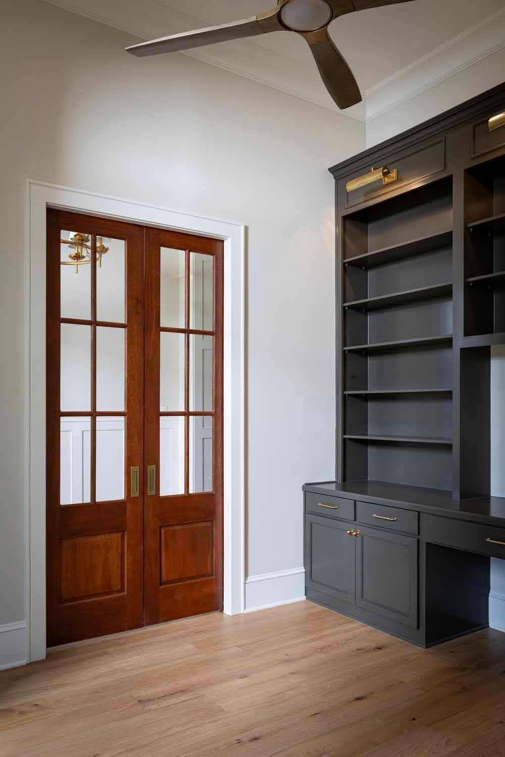 A wooden double French door with glass panels beside a dark grey built-in desk and bookshelf in a room with wood floors.