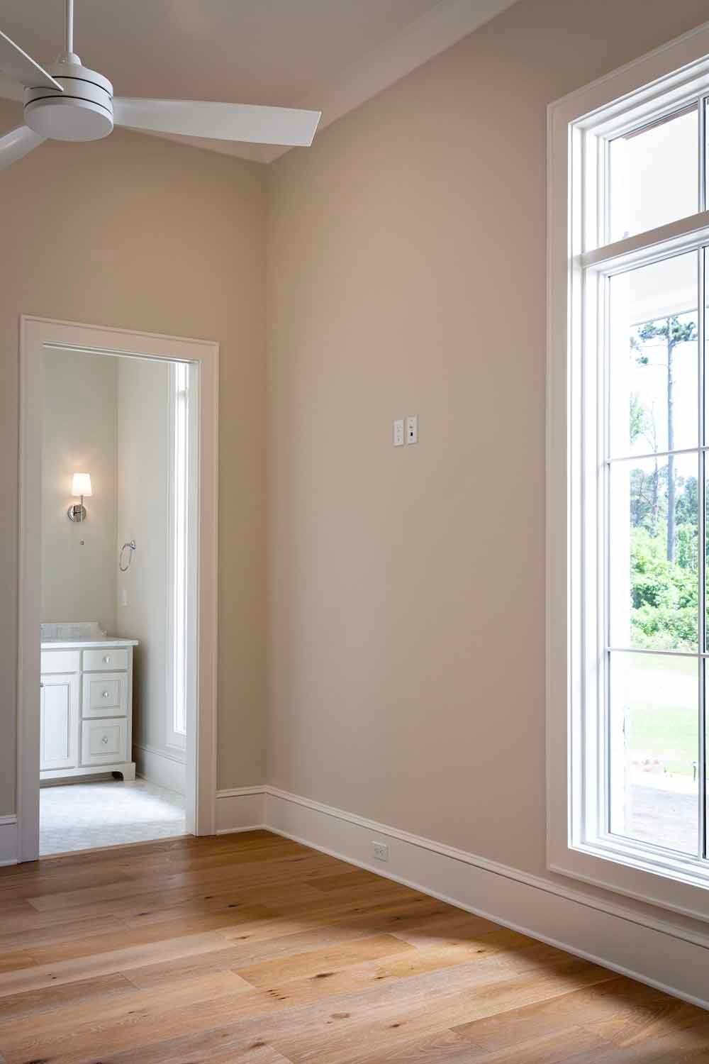 A bright room with light beige walls, hardwood floors, a white ceiling fan, and a view into a bathroom through a doorway.