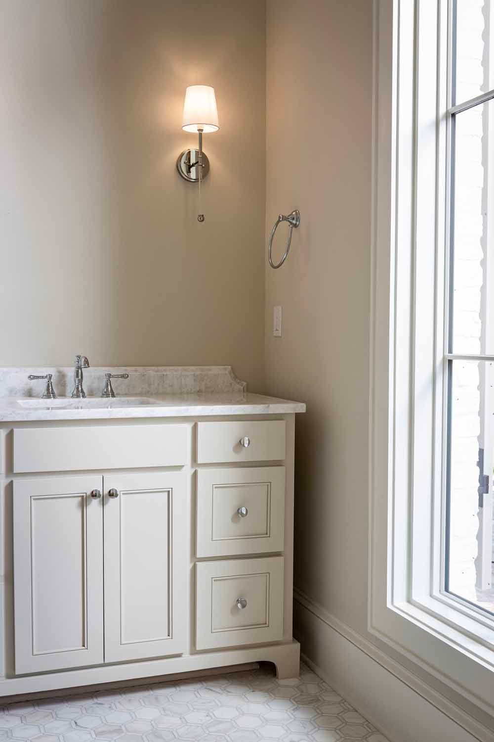A bathroom vanity with a white cabinet, marble countertop, silver faucet, and a wall-mounted light fixture near a window.