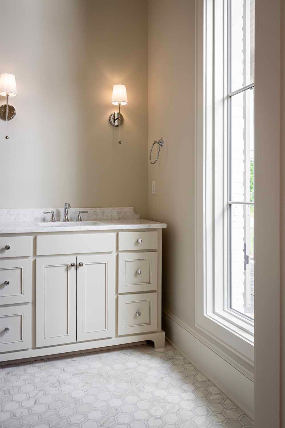 A white bathroom vanity with marble top, two wall sconces, and a tall window in a room with neutral walls and tile floors.