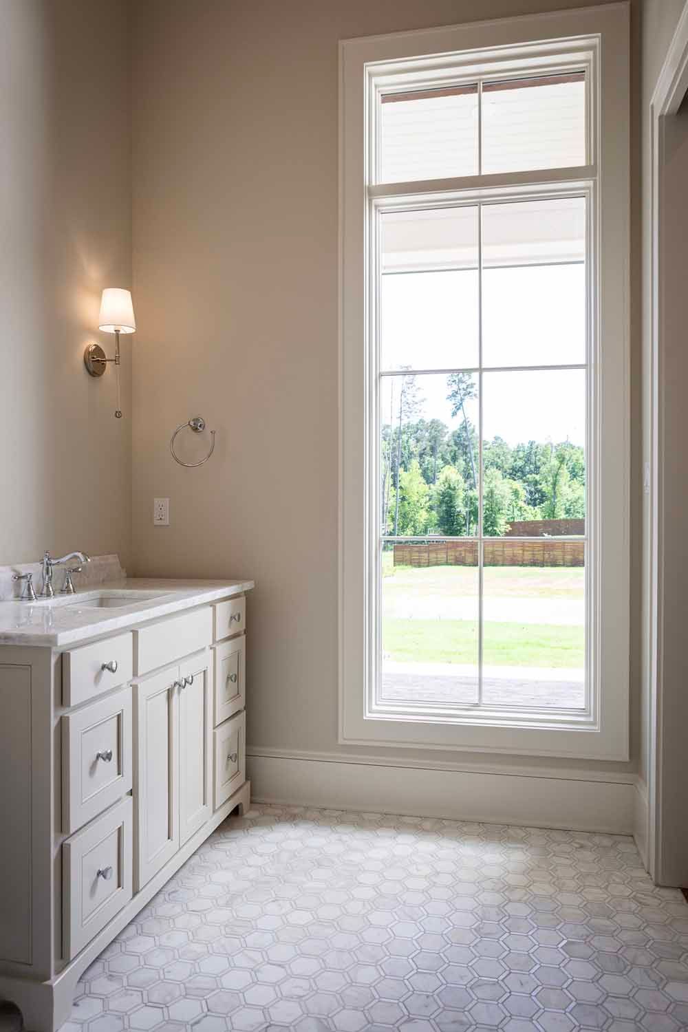 A light-colored vanity sits next to a large floor-to-ceiling window in a bright bathroom with tiled floors.