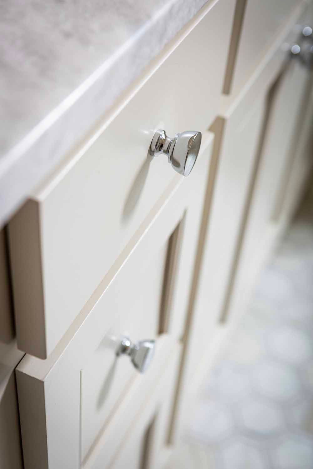 Close-up of white bathroom cabinets with chrome knobs and a stone countertop, angled view.