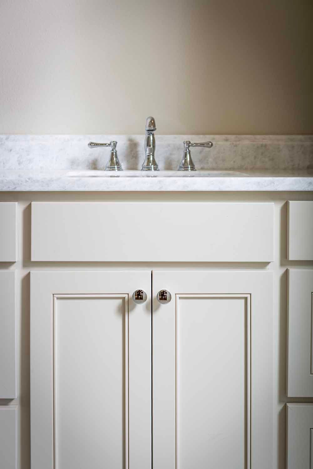 A close-up of a white vanity cabinet with a marble countertop, undermount sink, and silver faucet handles.