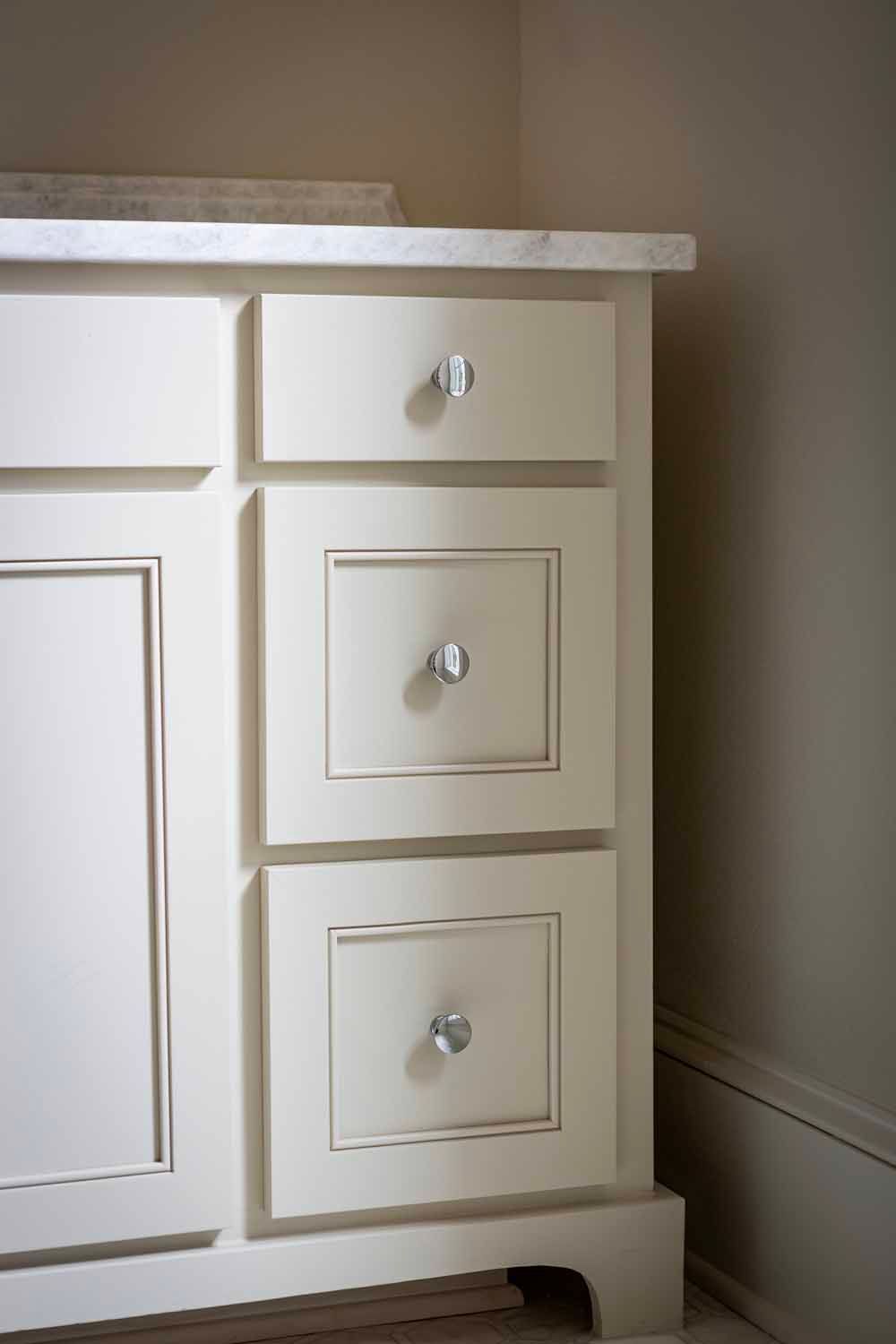 A cream-colored bathroom vanity cabinet with three drawers, chrome knobs, and a white countertop.