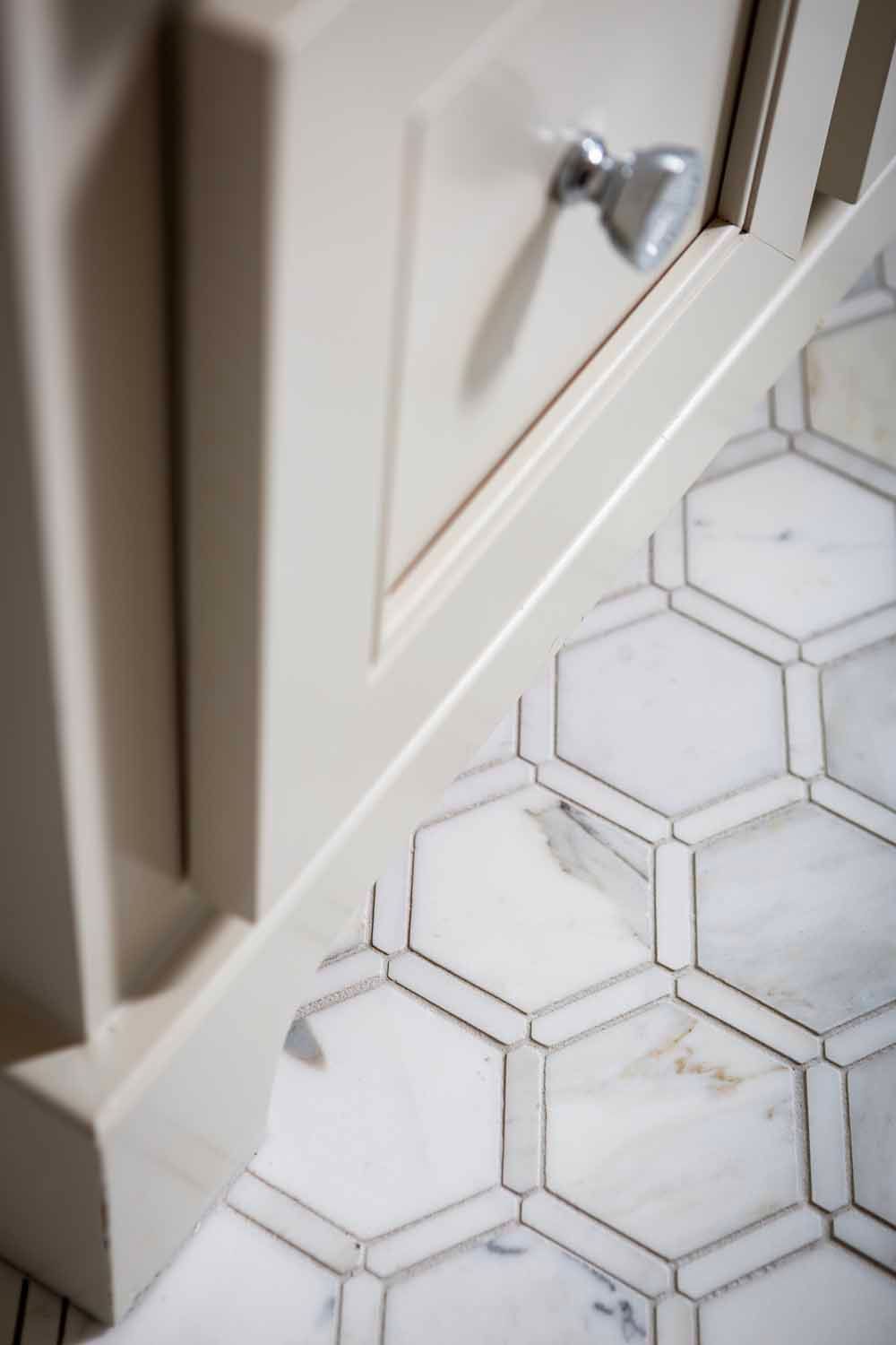 A close-up of a cream-colored cabinet vanity with a silver knob, positioned over a white marble hexagonal tile floor.
