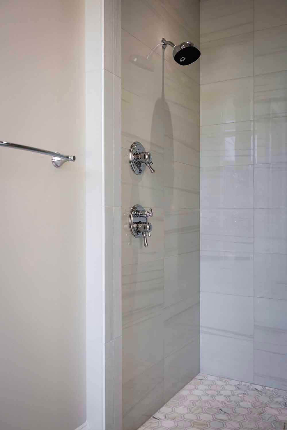 A modern walk-in shower with light tile walls, a circular stone floor, and two chrome faucet controls.
