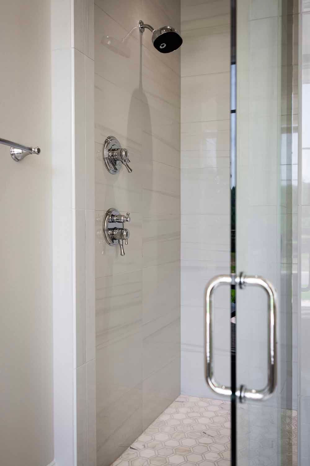 A walk-in shower with a glass door, chrome fixtures, light-colored tiled walls, and a pebble-tiled floor.