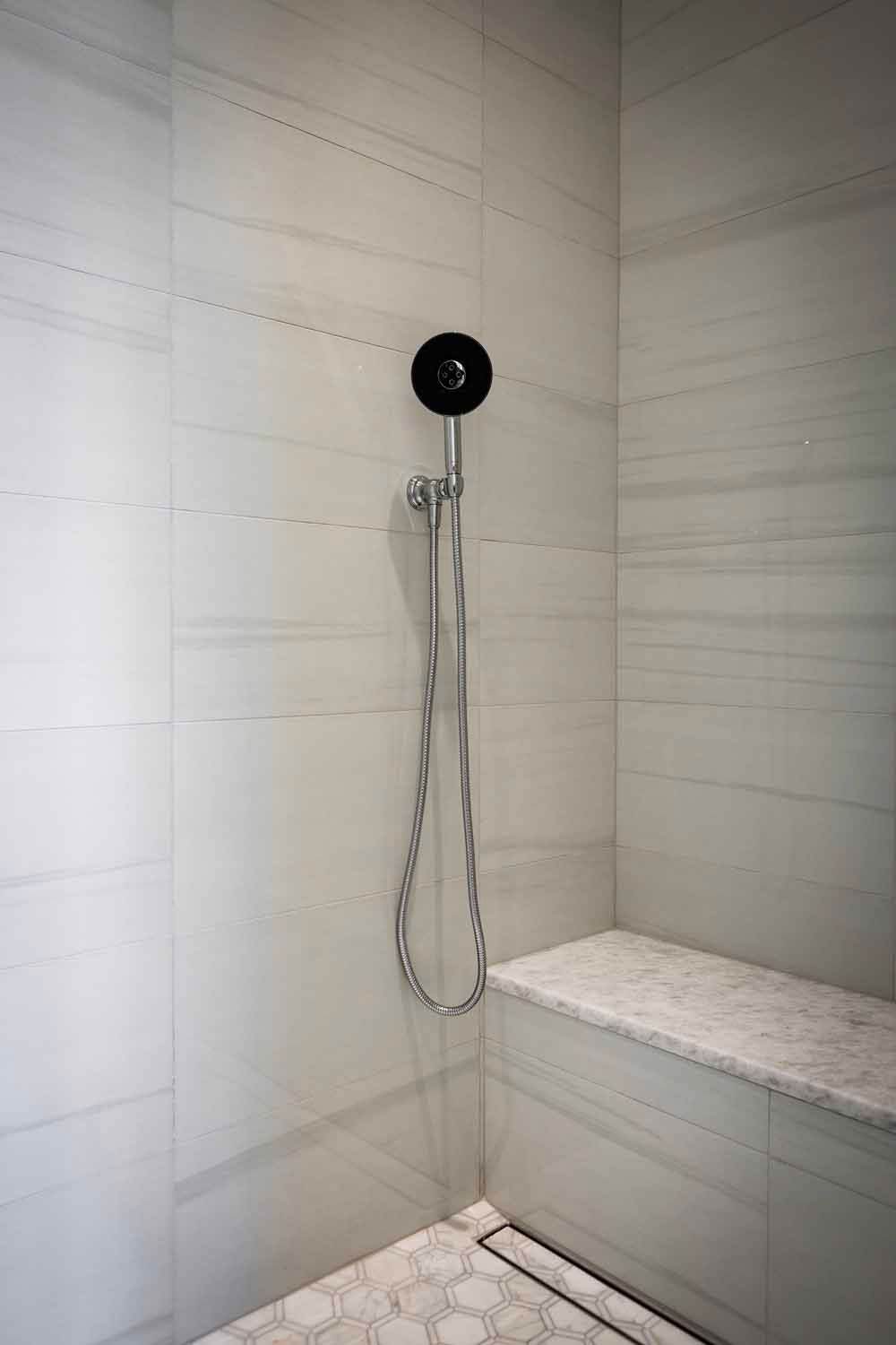 A modern tiled shower with a black handheld showerhead, a stone bench, and a linear floor drain.