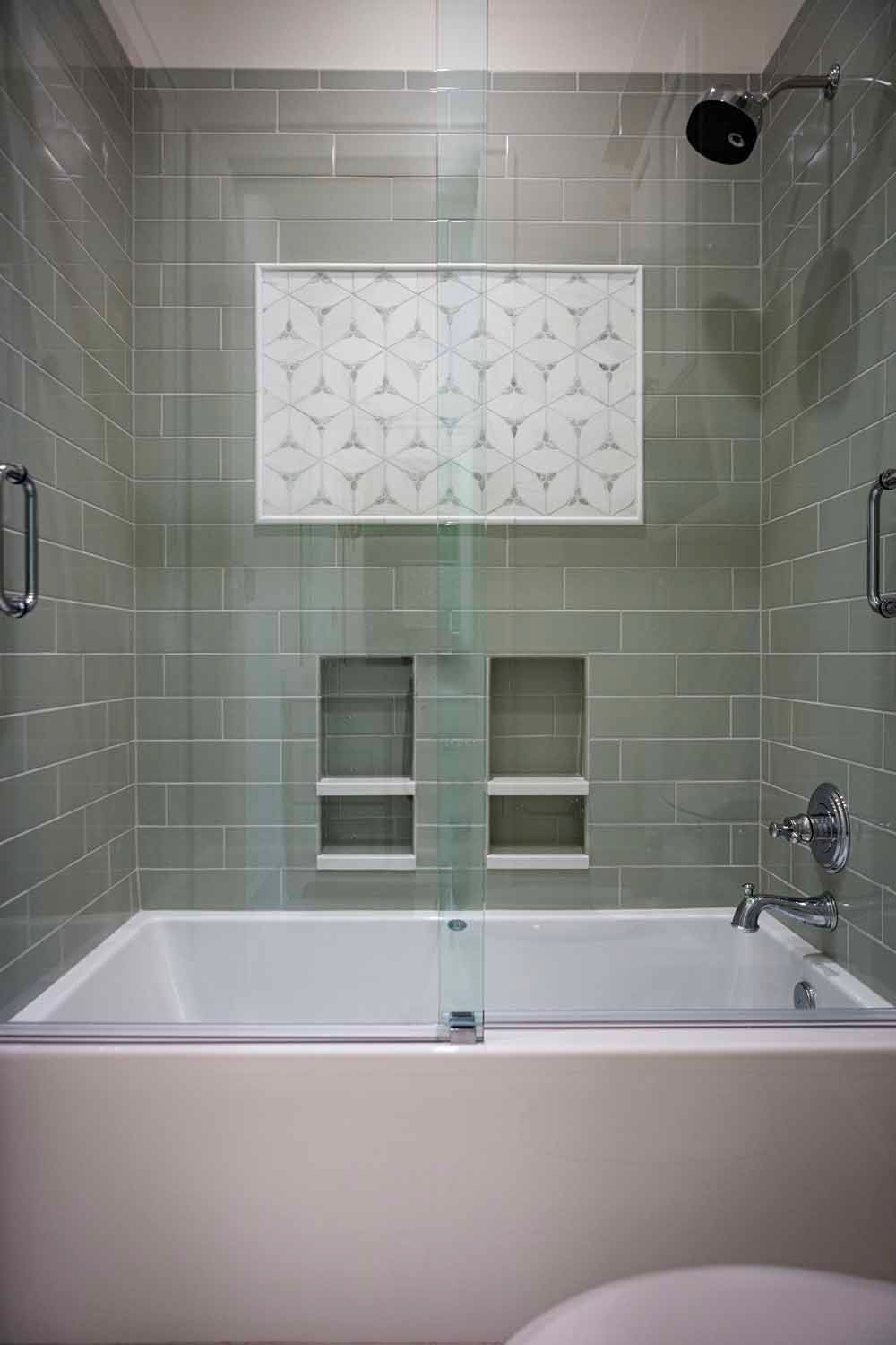 A bathtub and shower combo with sage subway tiles, dual wall niches, and a decorative patterned accent tile.