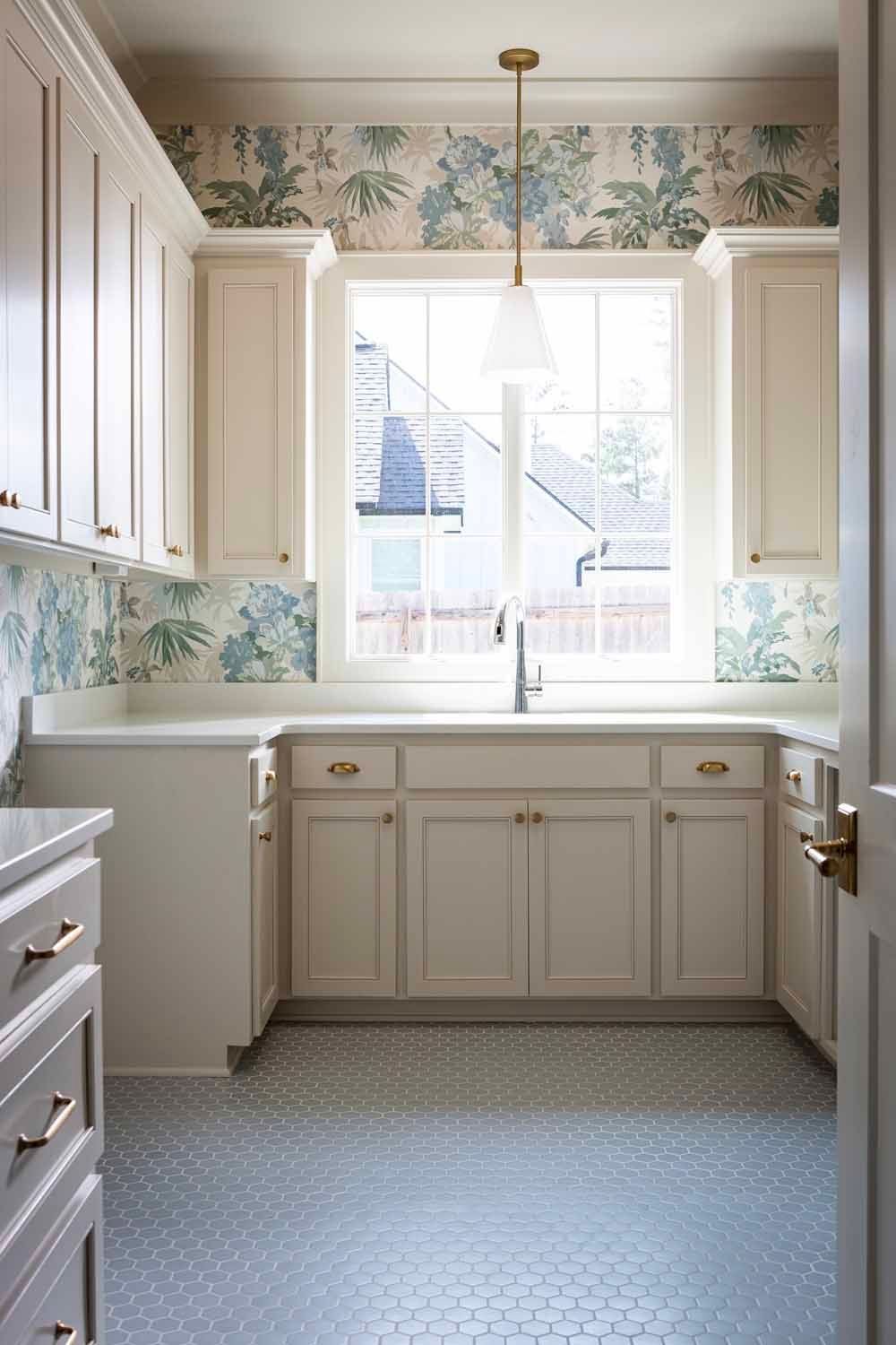 A bright, elegant kitchen with cream cabinets, blue and green floral wallpaper, a central sink, and hexagon floor tiles.