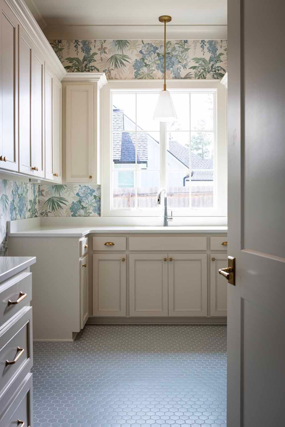A bright kitchen with off-white cabinets, blue floral wallpaper, a central window with a sink, and light gray tile flooring.