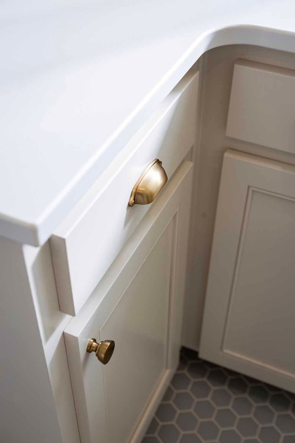Cream-colored kitchen cabinets with brass hardware and a white countertop over a grey hexagonal tile floor.
