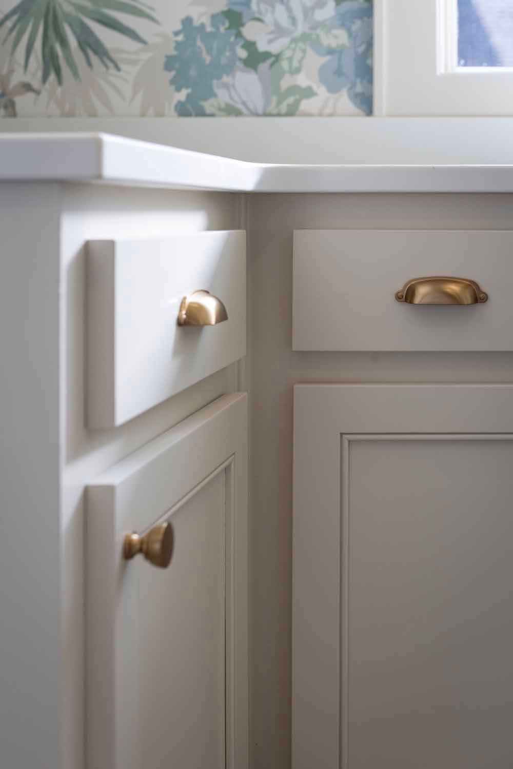 Off-white kitchen cabinets with gold cup pulls and a knob, set against a floral wallpaper backsplash.