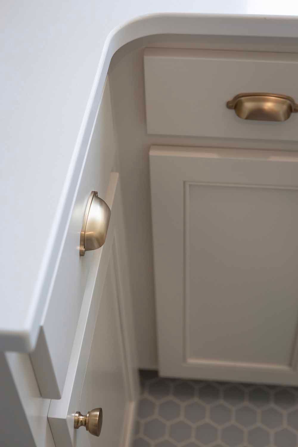Cream-colored kitchen cabinets with brass cup pulls and knobs beneath a rounded countertop above a grey hexagon tile floor.