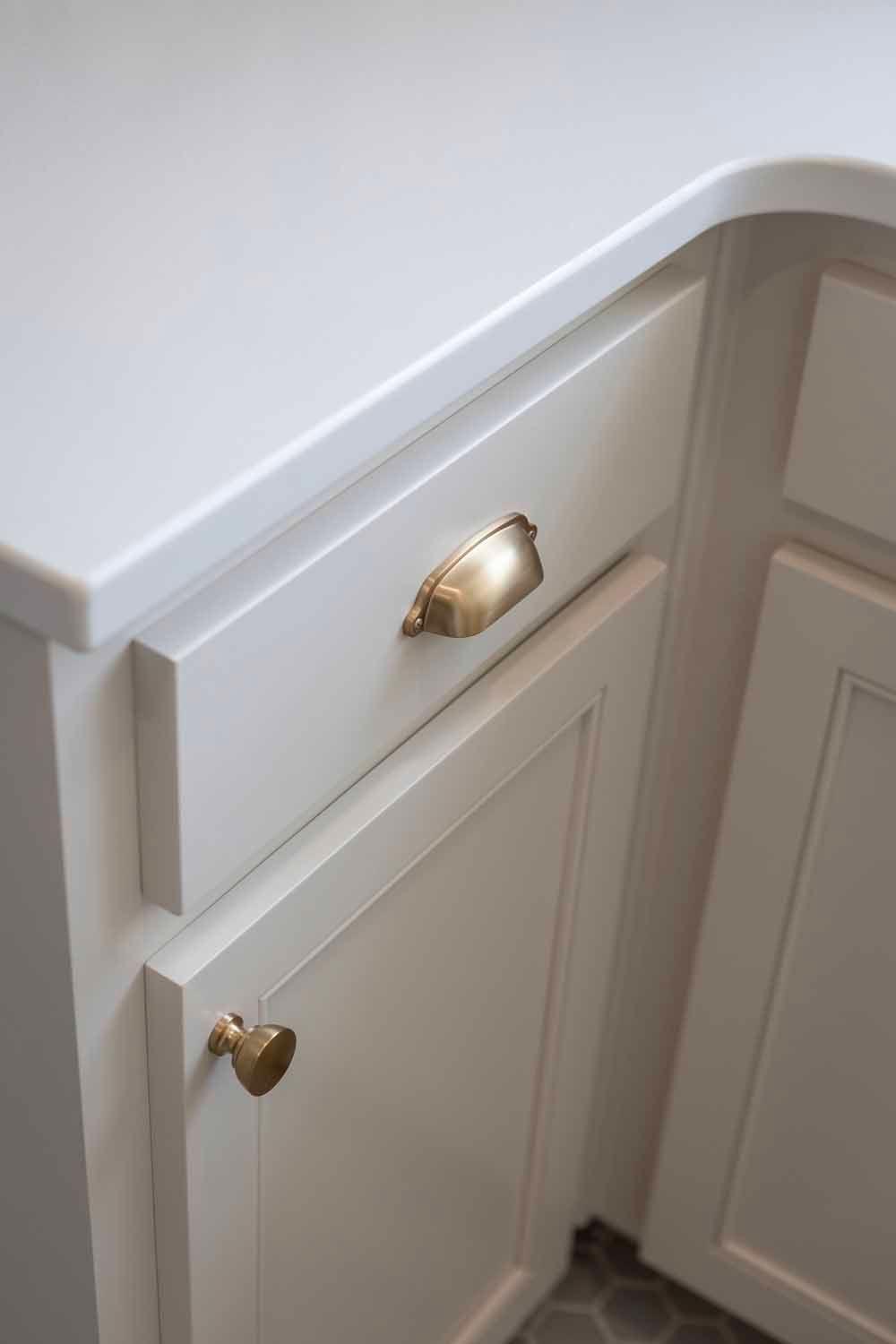 A close-up of a white kitchen cabinet with brass cup pulls and a round knob, featuring a smooth, light-colored countertop.