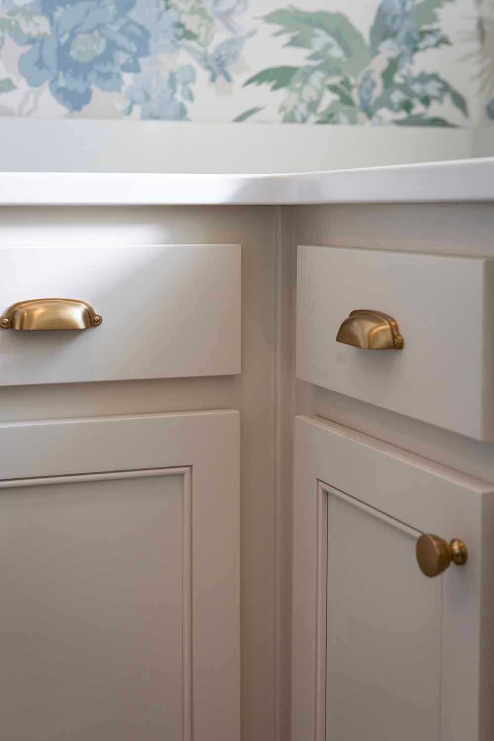 Cream-colored kitchen cabinets with brass cup pulls and a knob, set against floral wallpaper.