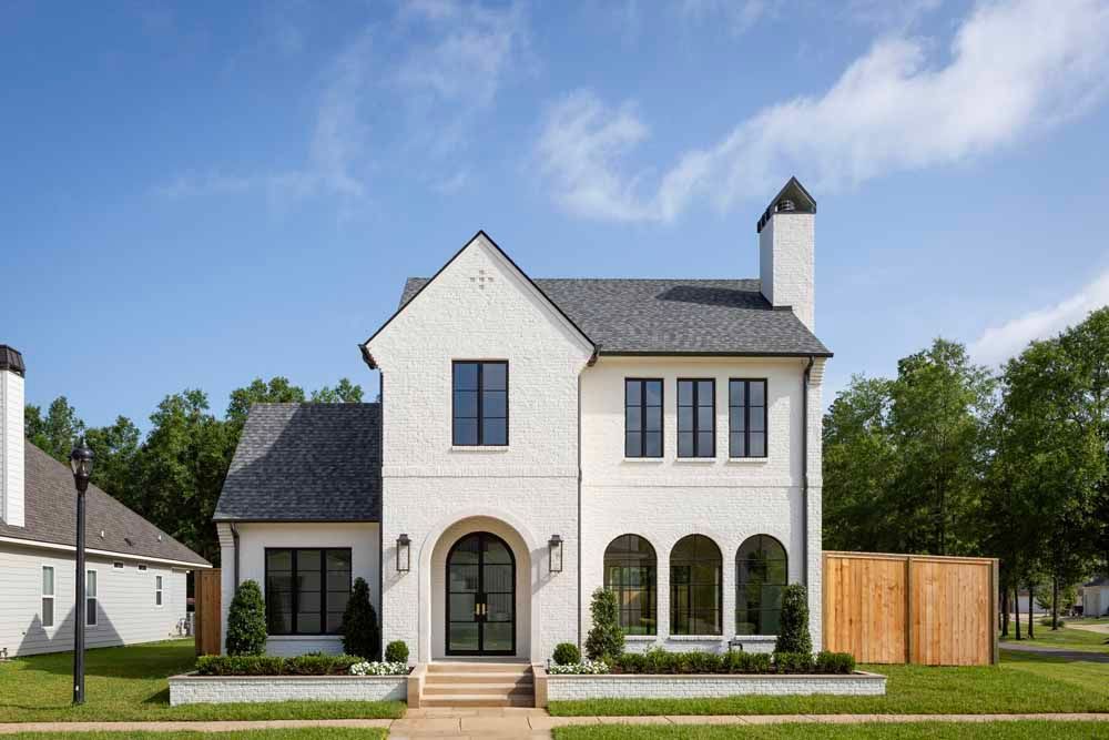 Two-story white brick house with black-trimmed windows, arched front entryway, and a grey shingled roof under a blue sky.