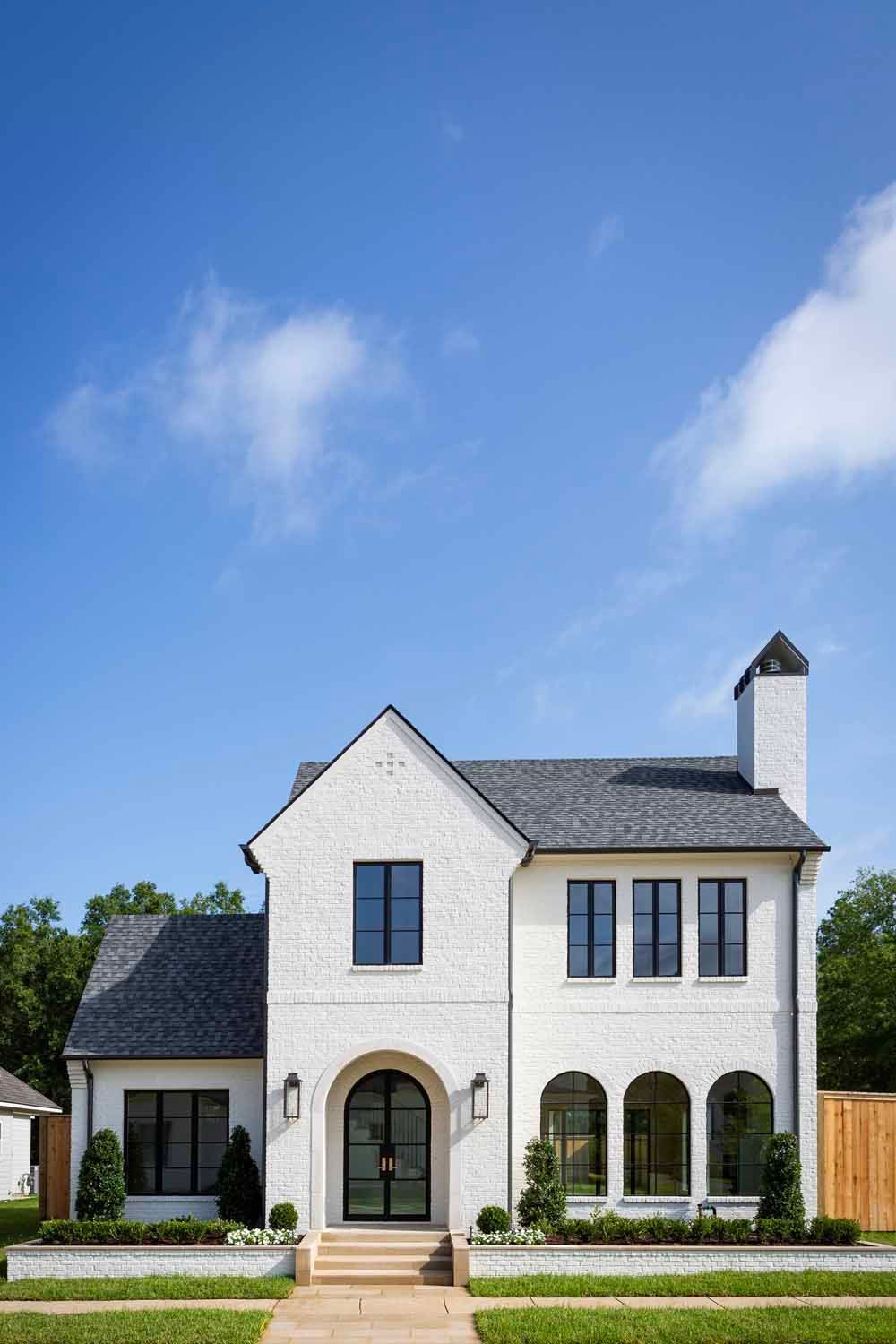 A two-story white brick house with a dark gabled roof, arched front doorway, and large windows under a clear blue sky.