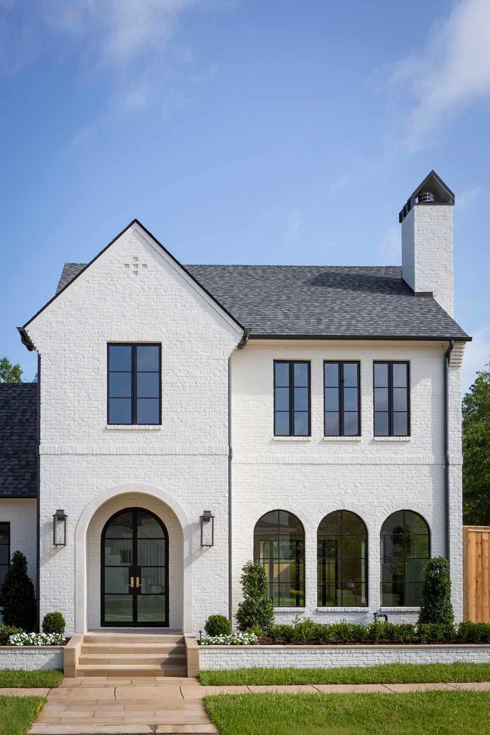 Two-story, white brick suburban house with a dark gabled roof, arched front door, and symmetrical windows under a blue sky.