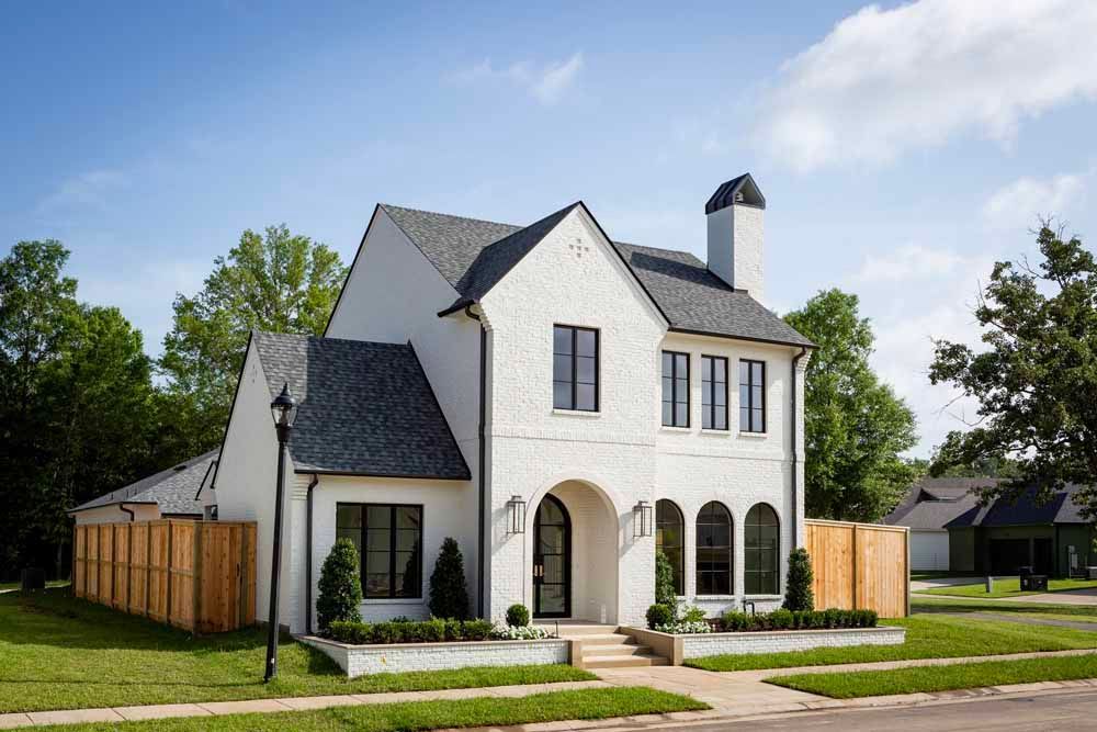 A two-story white brick house with a dark gabled roof, an arched entryway, and surrounding wooden fencing on a sunny day.