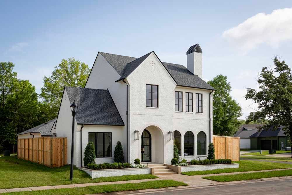 A two-story white brick house with a steep gray roof, arched entryway, and large windows set on a grassy suburban street.