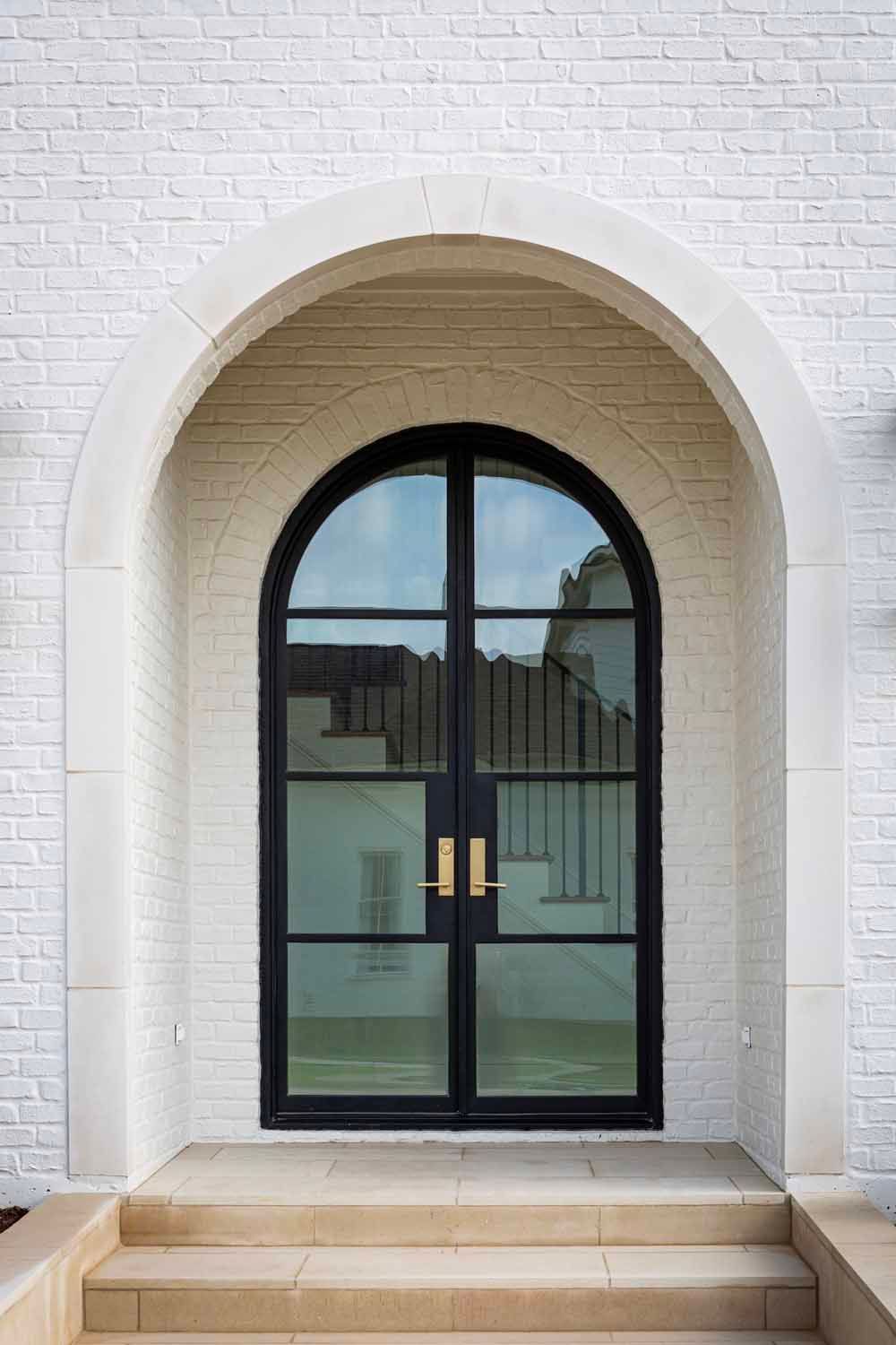 Arched double doors with black metal frames and brass hardware, set in a white brick wall above stone steps.