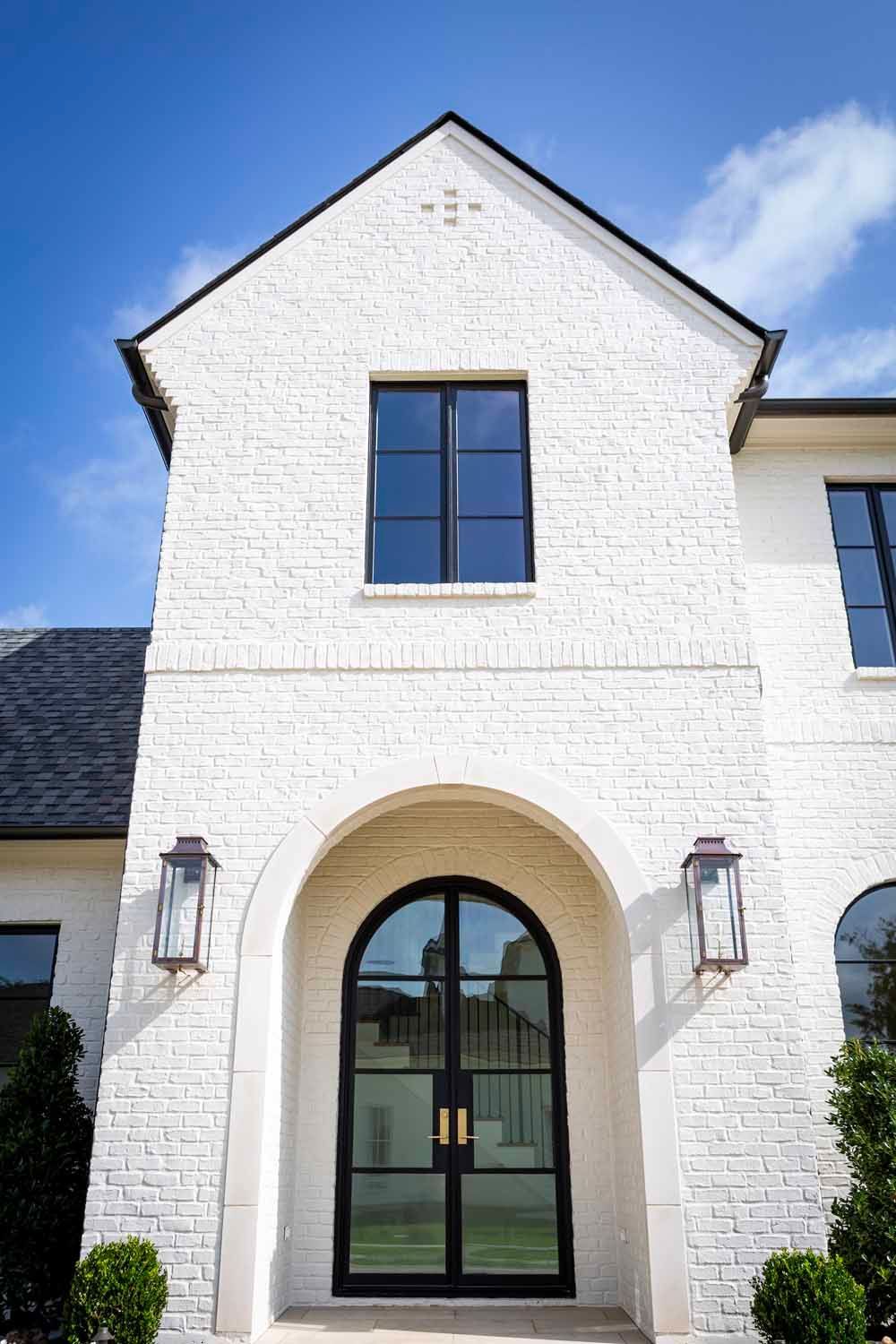 White brick home facade with a centered arched double door entry and dark-framed windows under a clear blue sky.