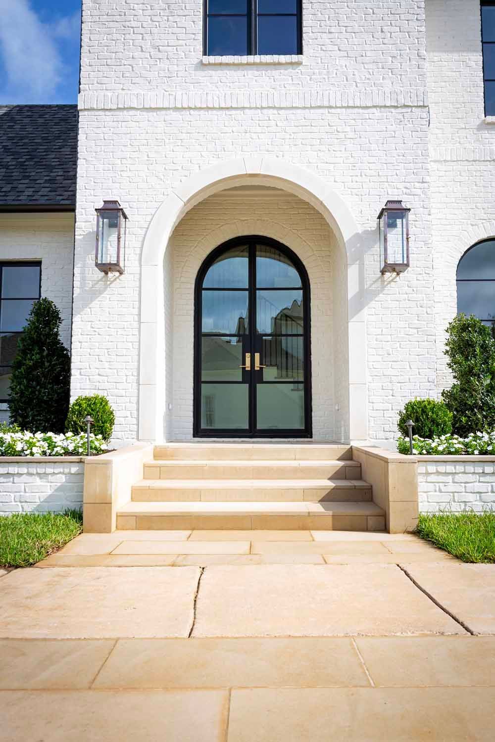 A white brick house exterior featuring a grand arched black metal double door, stone steps, and symmetrical lanterns.