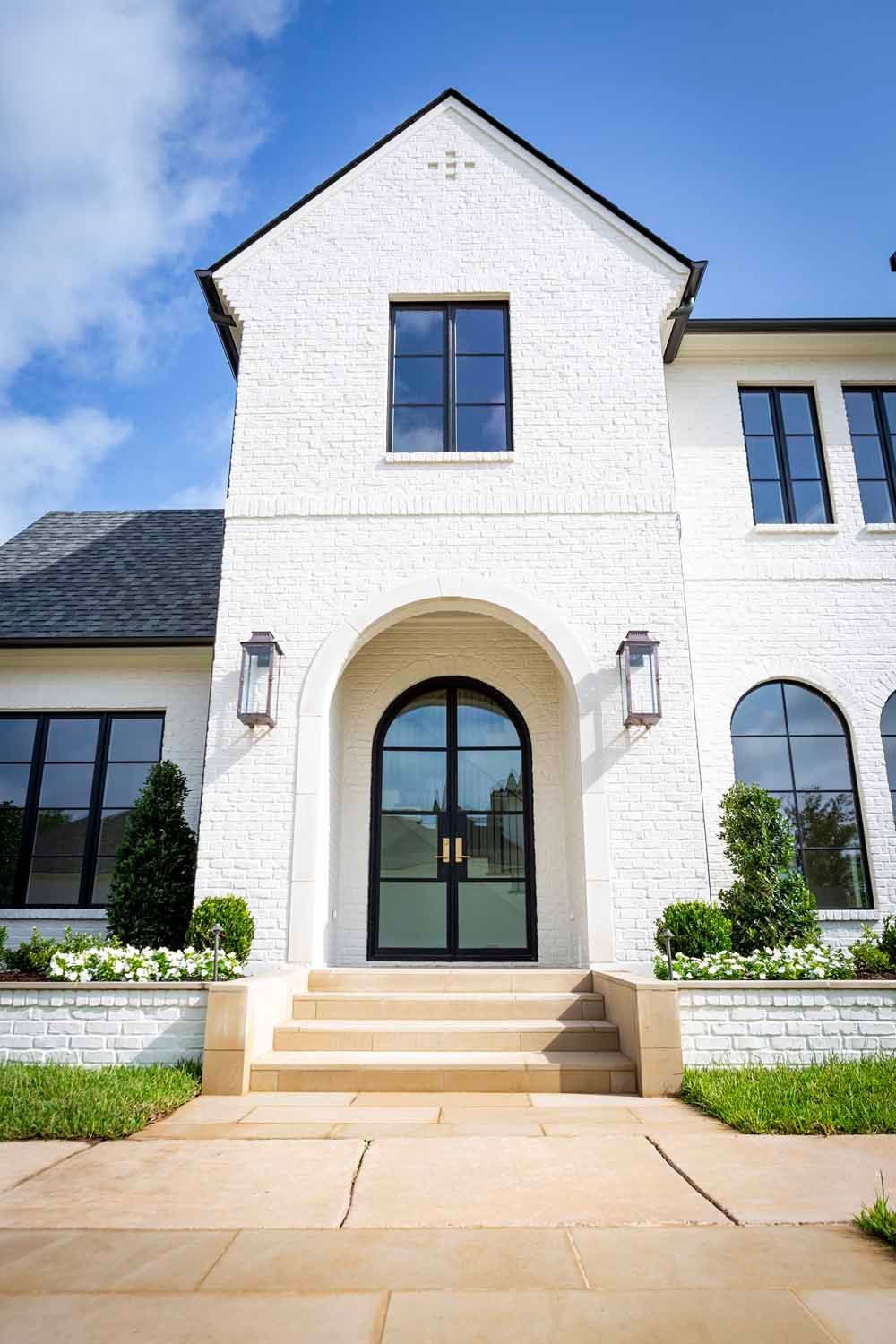 A white brick house with an arched entryway, black-framed windows, and stone steps leading to a modern black glass door.
