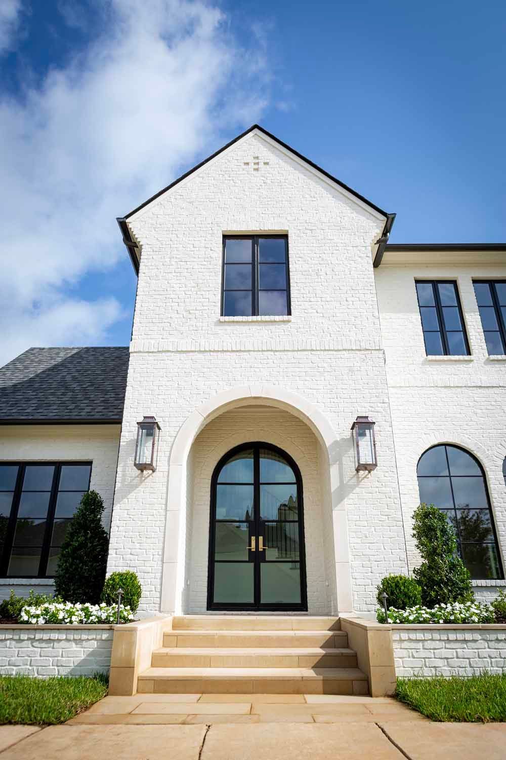 A white brick house with a gabled roof, an arched black-framed double door, and stone steps under a bright blue sky.