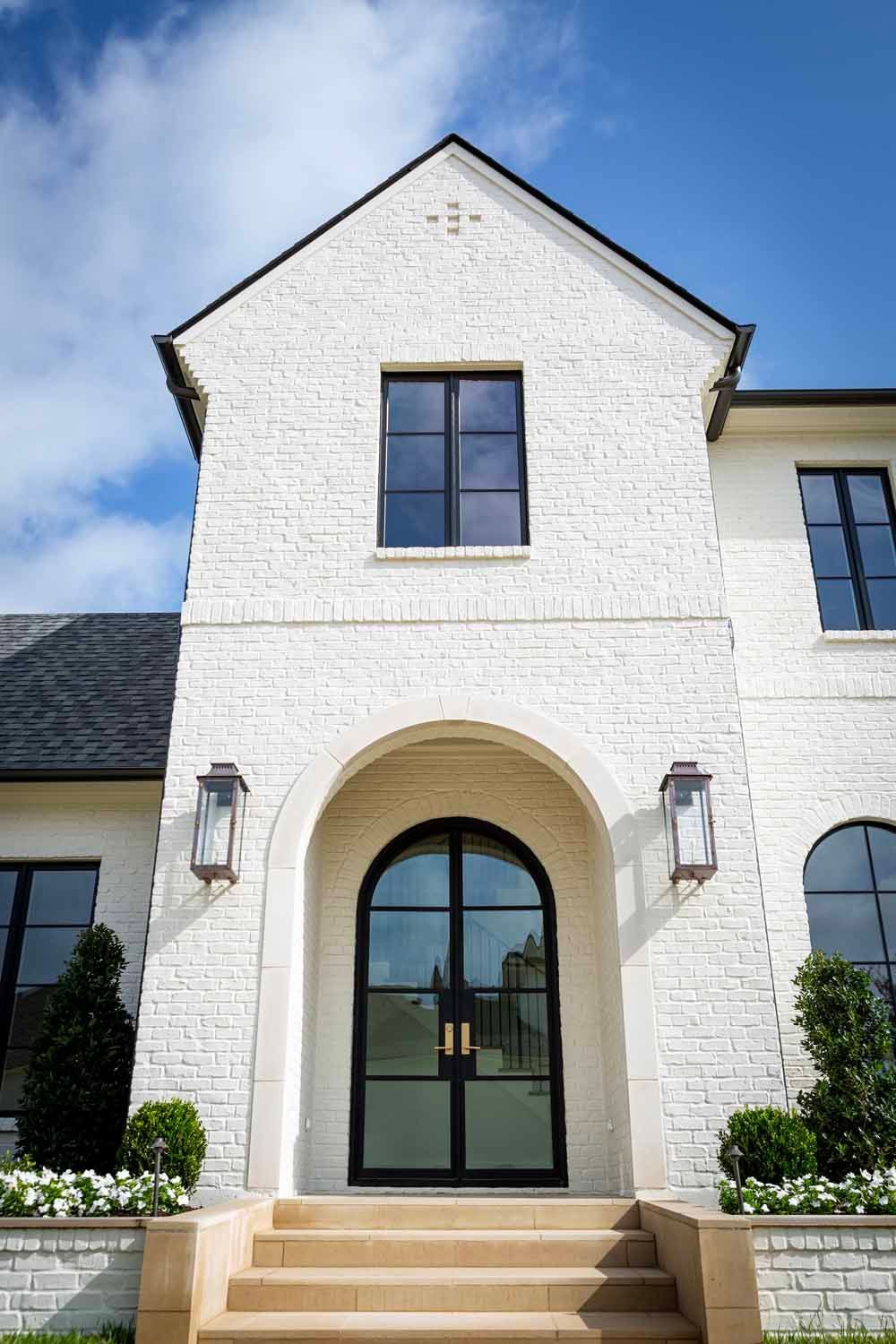 A white brick home entrance with an arched doorway, dark-framed windows, and stone steps under a bright blue sky.