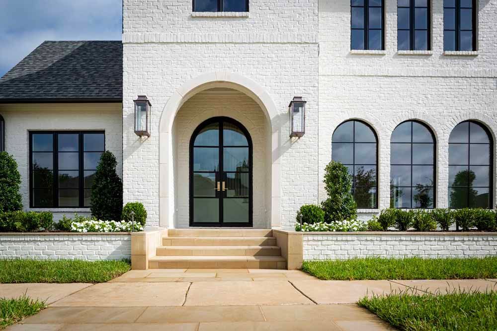 Front view of a white brick home with black-framed arched windows, a double arched door, and front stone steps.