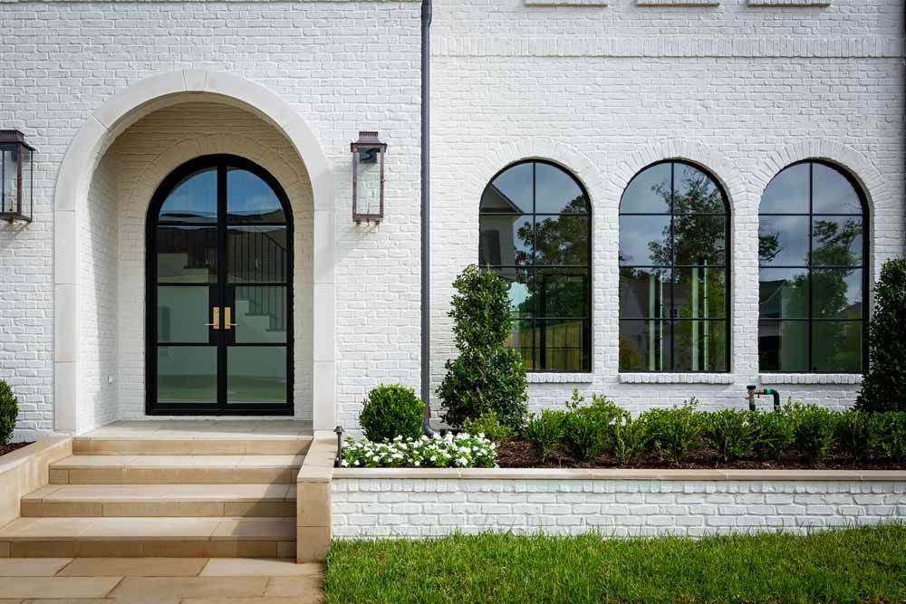 A white brick house exterior with a large arched double door, three arched windows, and a landscaped stone planter.