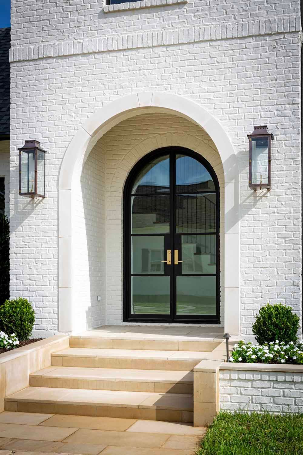 An arched, black-framed glass double door centered in a white brick exterior, flanked by lanterns and stone steps.