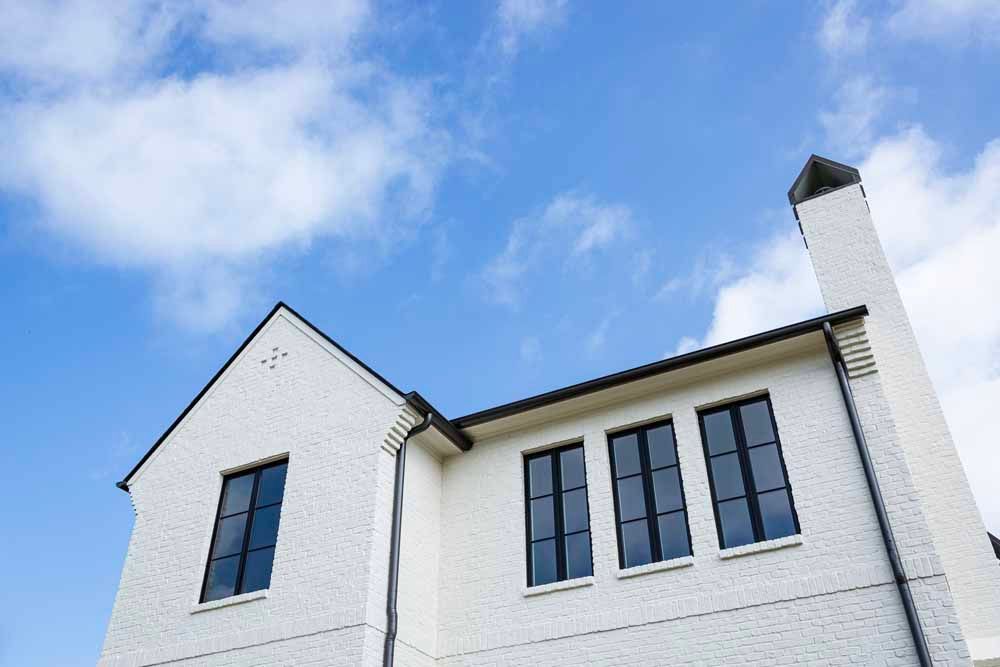 A low-angle view of a white brick building with dark-framed windows and a tall chimney against a bright blue cloudy sky.
