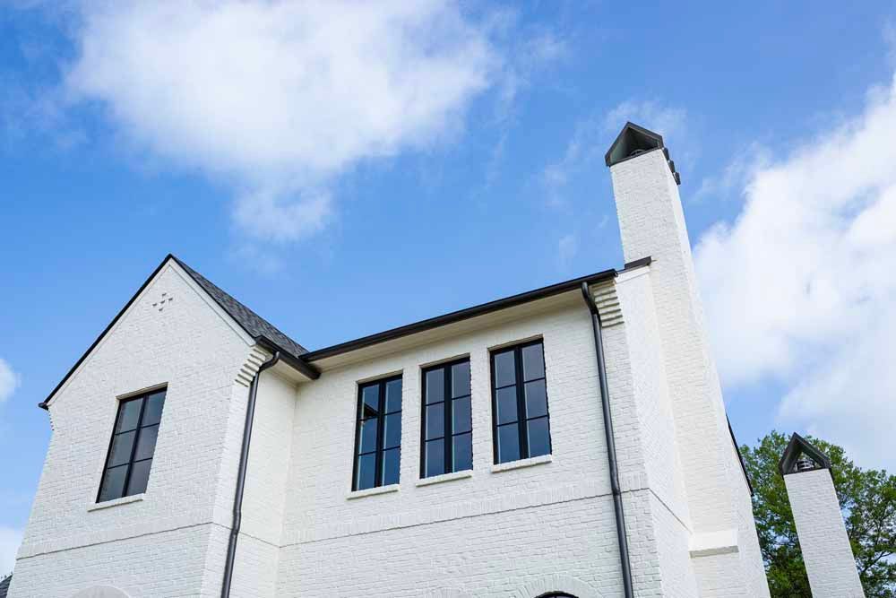 Low-angle view of a white brick building with black-framed windows and a prominent chimney against a bright blue sky.