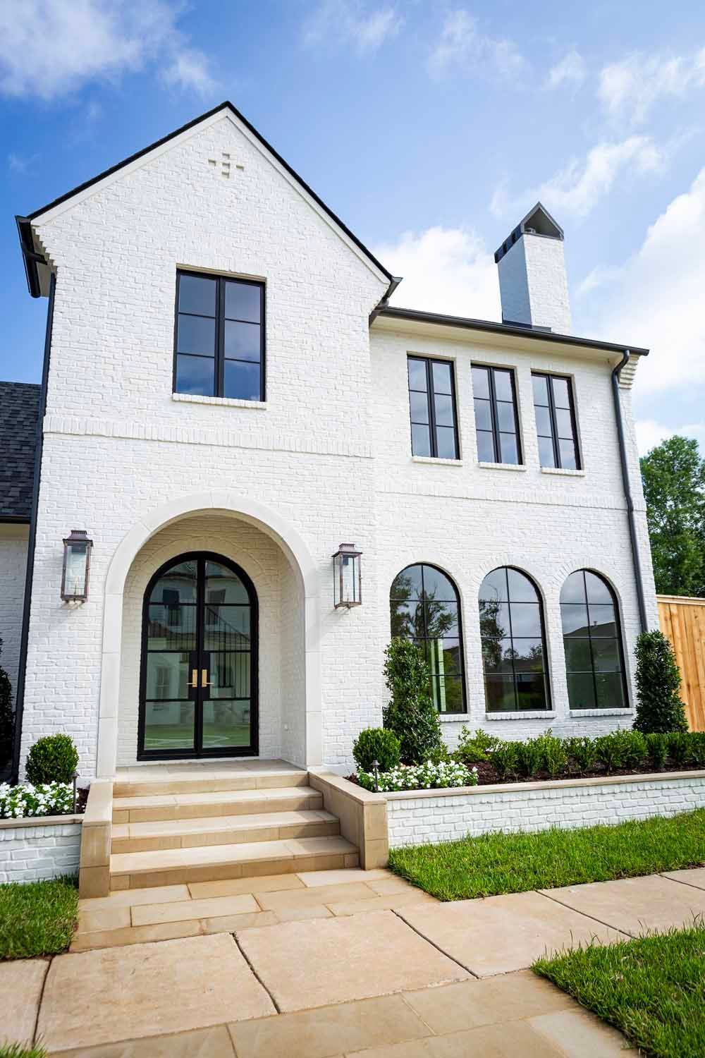 A white, two-story brick house with a front arched door, dark window frames, a chimney, and stone steps under a blue sky.