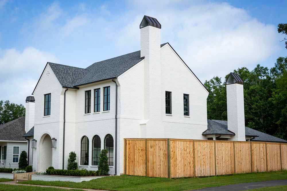 A two-story white brick house with dark shingled roofs, arched windows, and a tall wooden privacy fence in front.