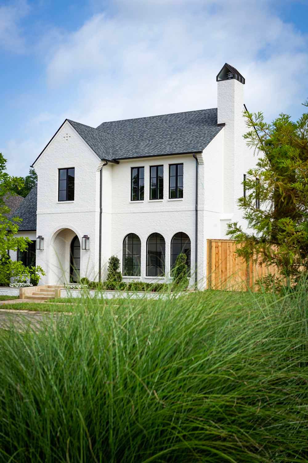 A two-story, white brick house with a dark gray shingled roof and arched windows, set behind tall green grasses.