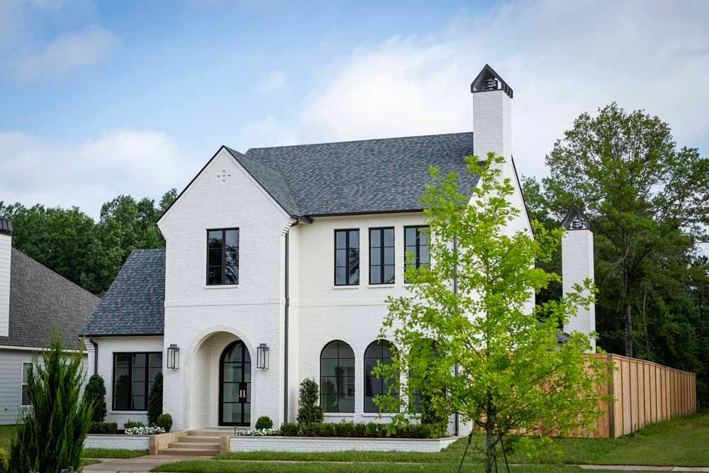 Two-story white brick house with a dark gabled roof, arched front entryway, and landscaped front yard under a blue sky.