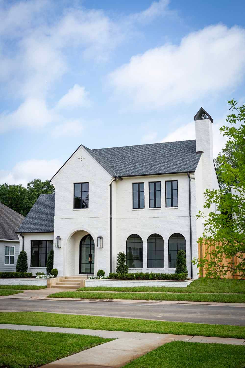 A two-story white brick house with a steep gray shingled roof, arched front entryway, and dark-framed windows.