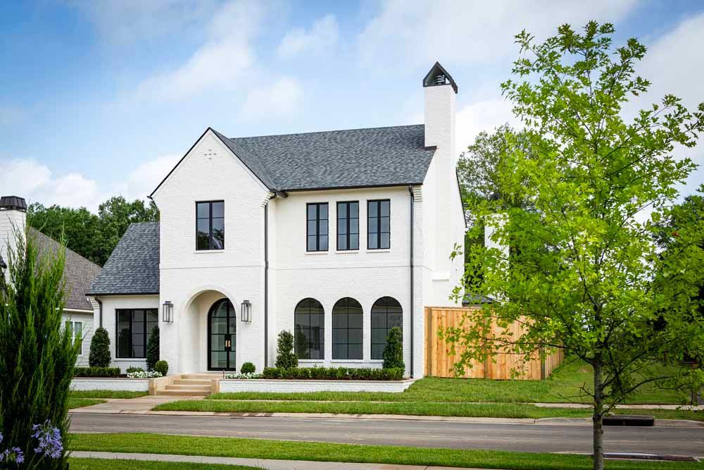 A two-story, white brick house with a dark gray shingled roof, black-framed windows, and a wooden fence on a sunny day.