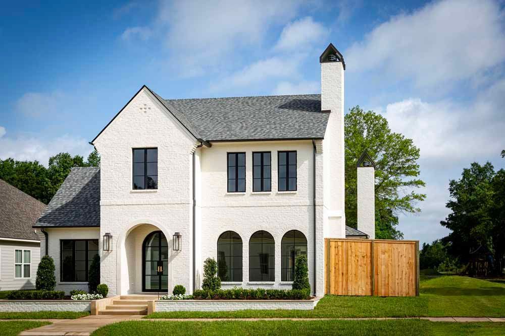 A two-story, white brick house with a dark gray shingled roof, arched entryway, and wooden privacy fence on a lawn.
