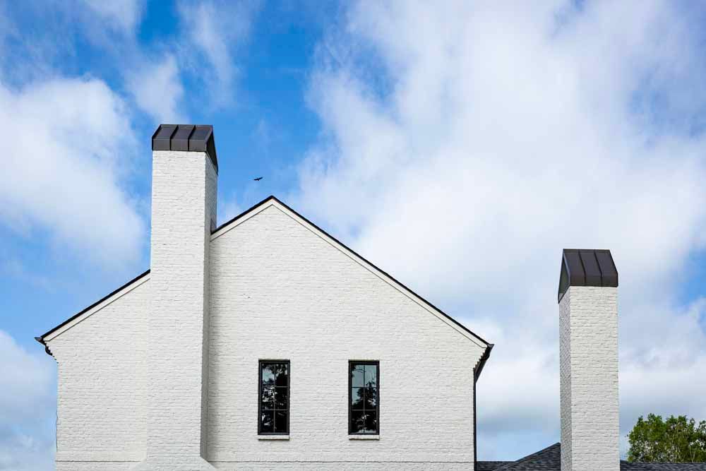 A white brick house exterior with two tall chimneys and black-framed windows against a blue, cloudy sky.