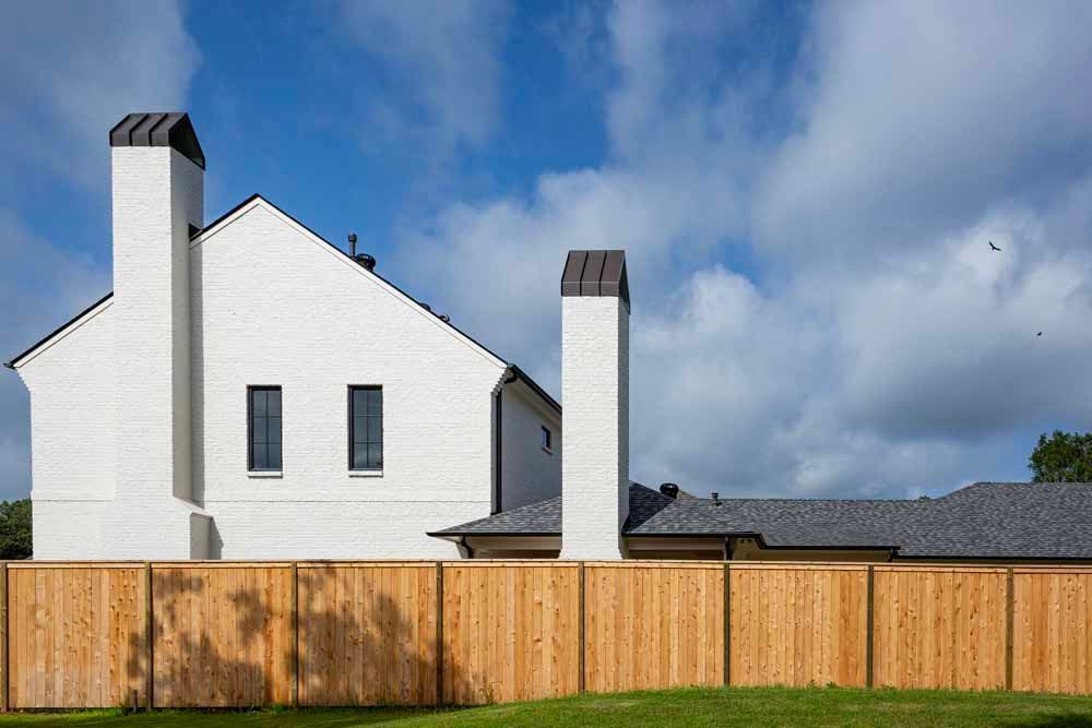 A white, two-story brick house with two tall chimneys and a gray shingled roof behind a tall wooden fence under a blue sky.
