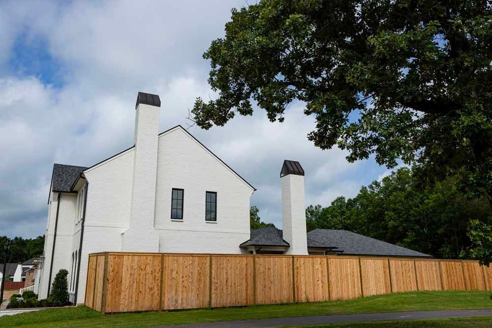 A white house with two prominent chimneys and a dark roof, enclosed by a new wooden privacy fence under a cloudy sky.