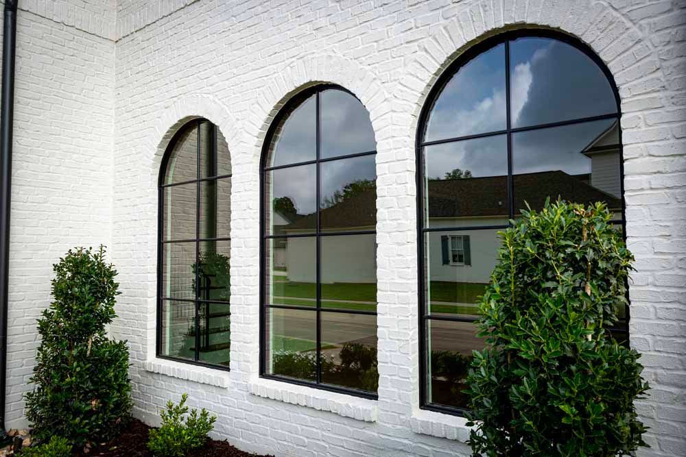 Three arched, black-framed windows on a white painted brick wall, flanked by green bushes.